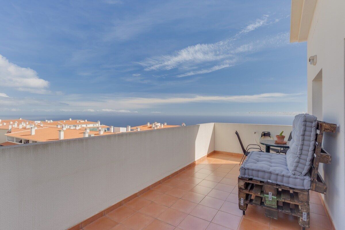 A balcony with a view of buildings, a cushioned chair, and a small table with a plant.