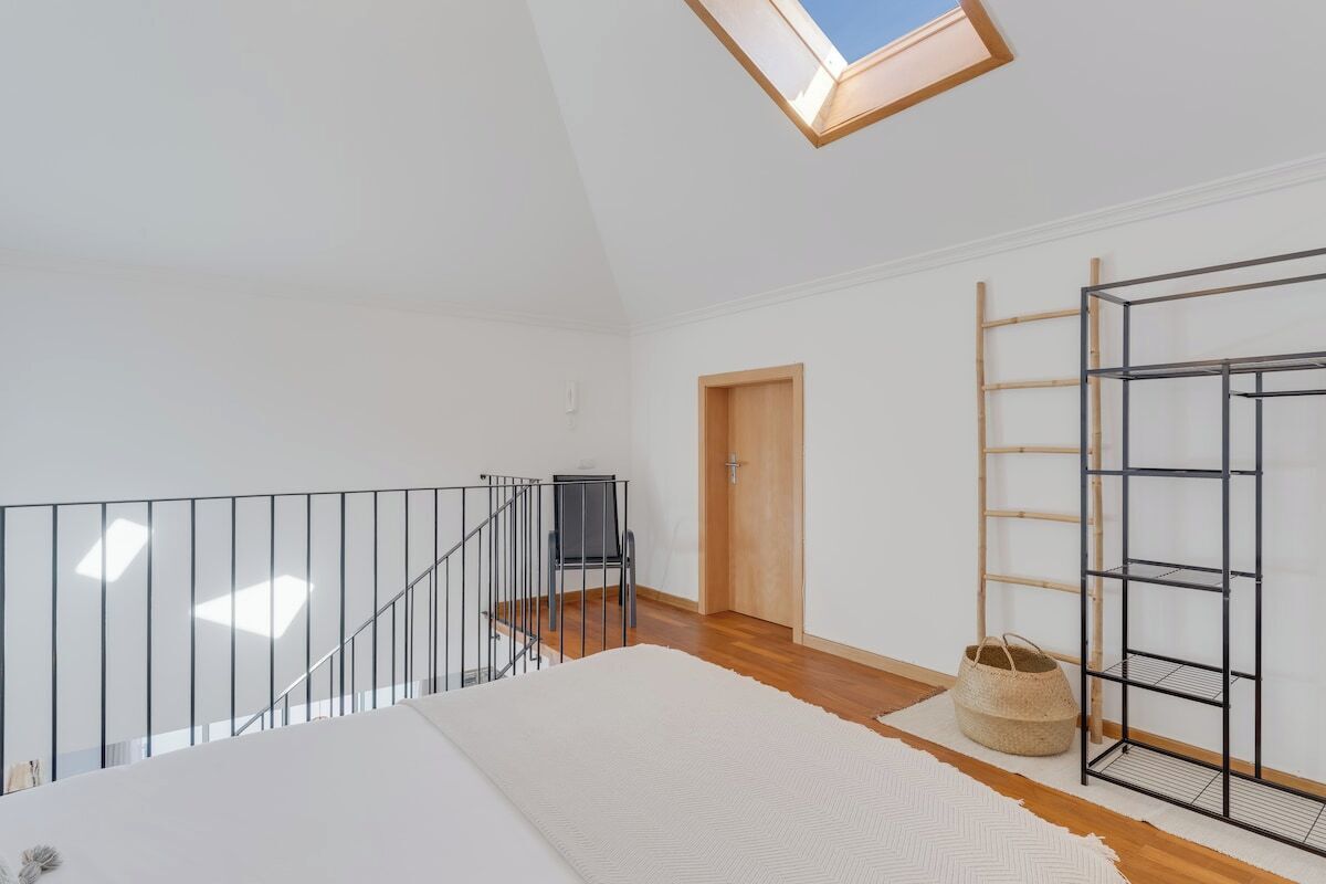 A spacious room with a wooden floor, a white wall, a skylight, a black metal staircase railing, and a black metal shelf unit.