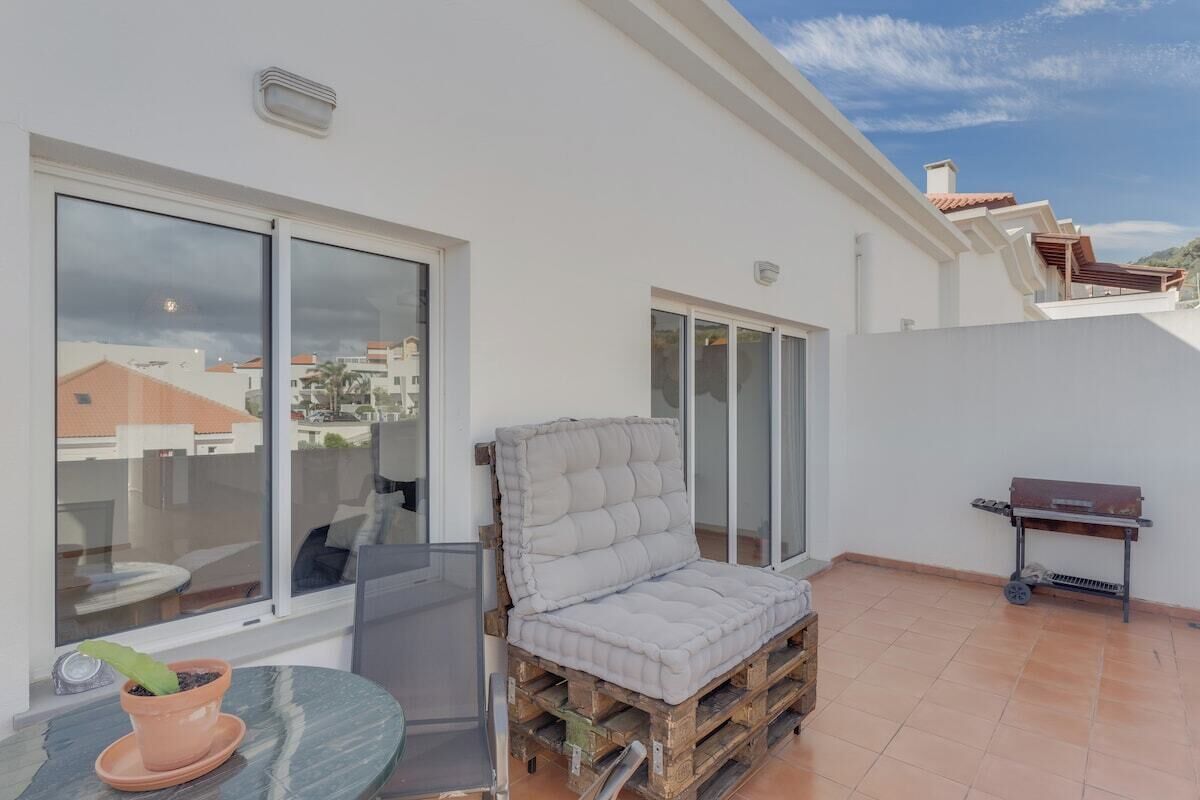 A balcony with a cushioned chair, a grill, and a potted plant.