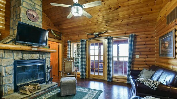 Ein Interieur einer Holzblockhütte mit einem Kamin aus Stein, einem darüber montierten Fernseher, einem Ledersofa und einem großen Fenster mit Vorhängen.