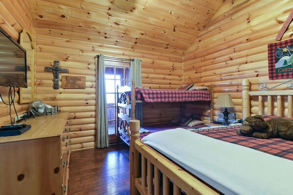 A log cabin bedroom with a wooden bed, a desk with a lamp, and a cross on the wall.