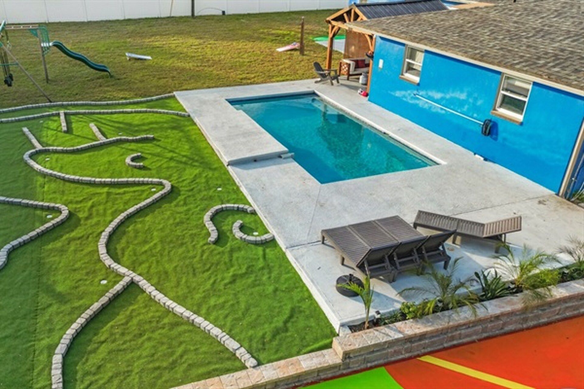 A swimming pool with a surrounding deck, a children's play area, and a blue building with windows.