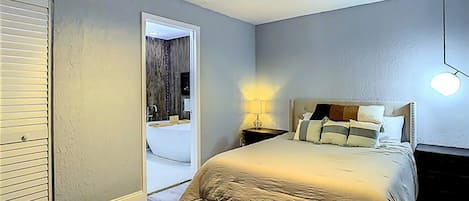 A bedroom with a bed, a nightstand, a lamp, a ceiling fan, and a door leading to a bathroom.