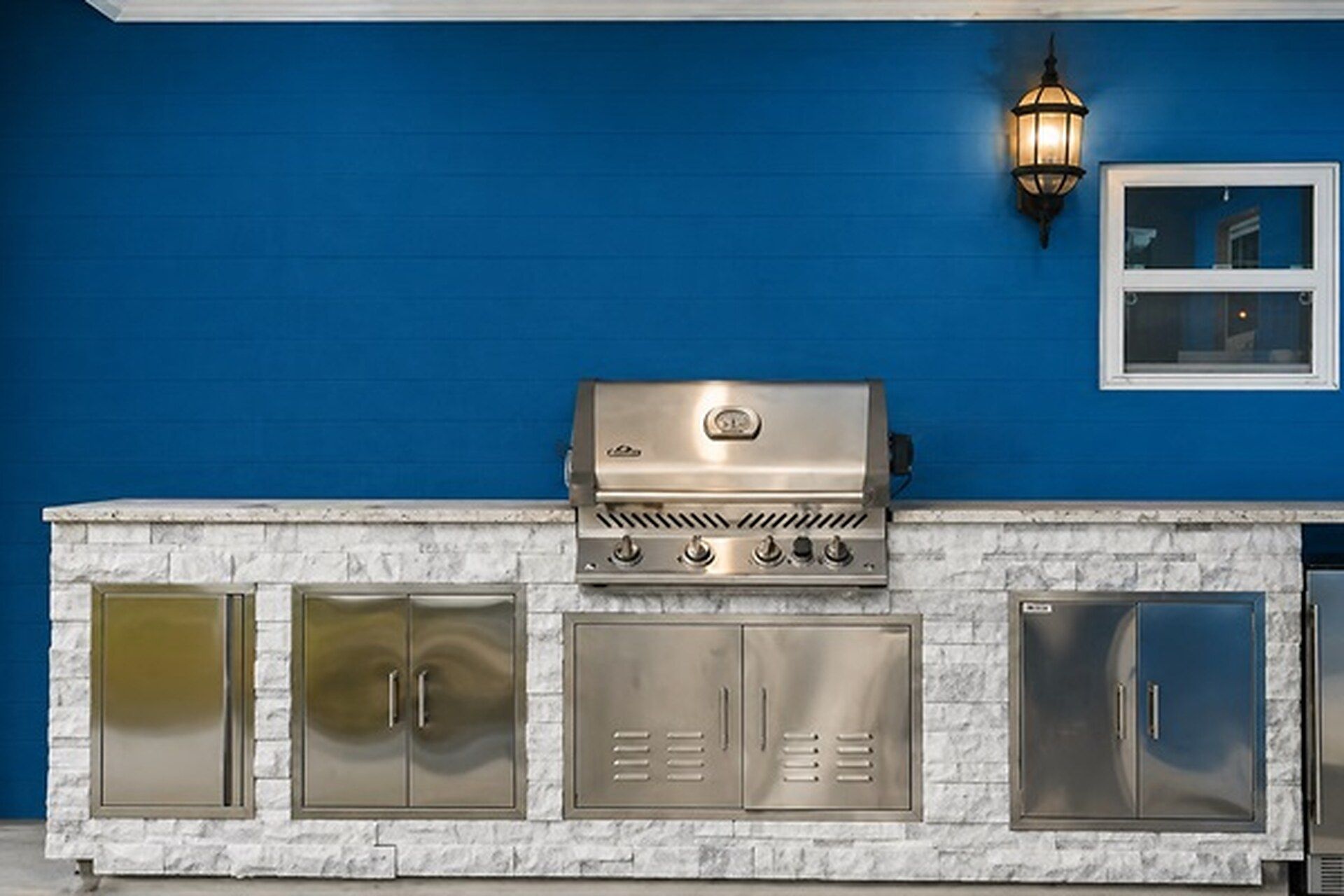 A barbecue grill with a stainless steel finish, a stone countertop, and a blue wall in the background.
