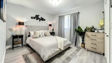 A modern bedroom with a large bed, bedside lamps, a dresser, and a potted plant.