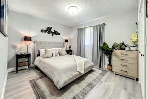 A modern bedroom with a large bed, bedside lamps, a dresser, and a potted plant.