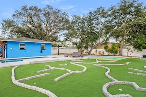 A backyard with a swimming pool, a blue house, and a lawn with a geometric pattern. - 2 Homes- Heated Pool & Pickleball- The Dunedin Duo (Dunedin, Florida)