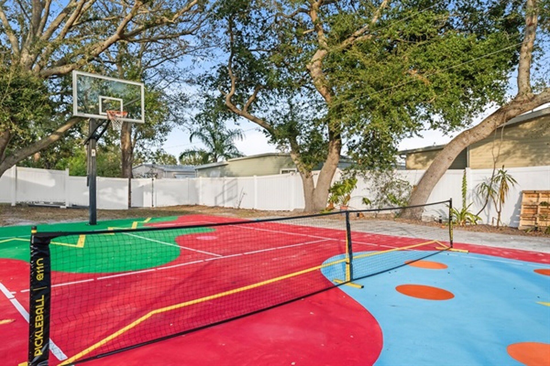 Un terrain de sport extérieur doté d’un panier de basketball, entouré d’arbres et d’une clôture blanche.
