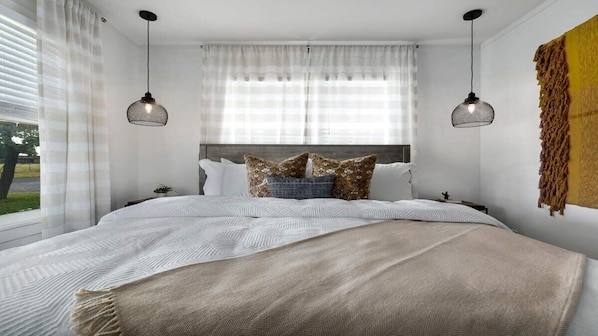 A modern bedroom with a large bed, two pendant lights, sheer curtains, and a view of the outdoors.
