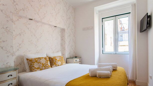Een slaapkamer met een bed, beddengoed in wit en geel, een houten vloer, een nachtkastje, een raam en een televisie aan de muur.