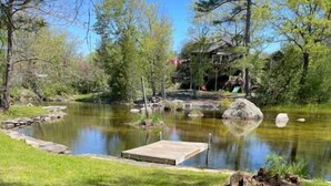 A serene pond with a wooden dock, surrounded by lush greenery and a rustic cabin in the background. - The Fisherman - 3-bedroom River-front Retreat (Selwyn)