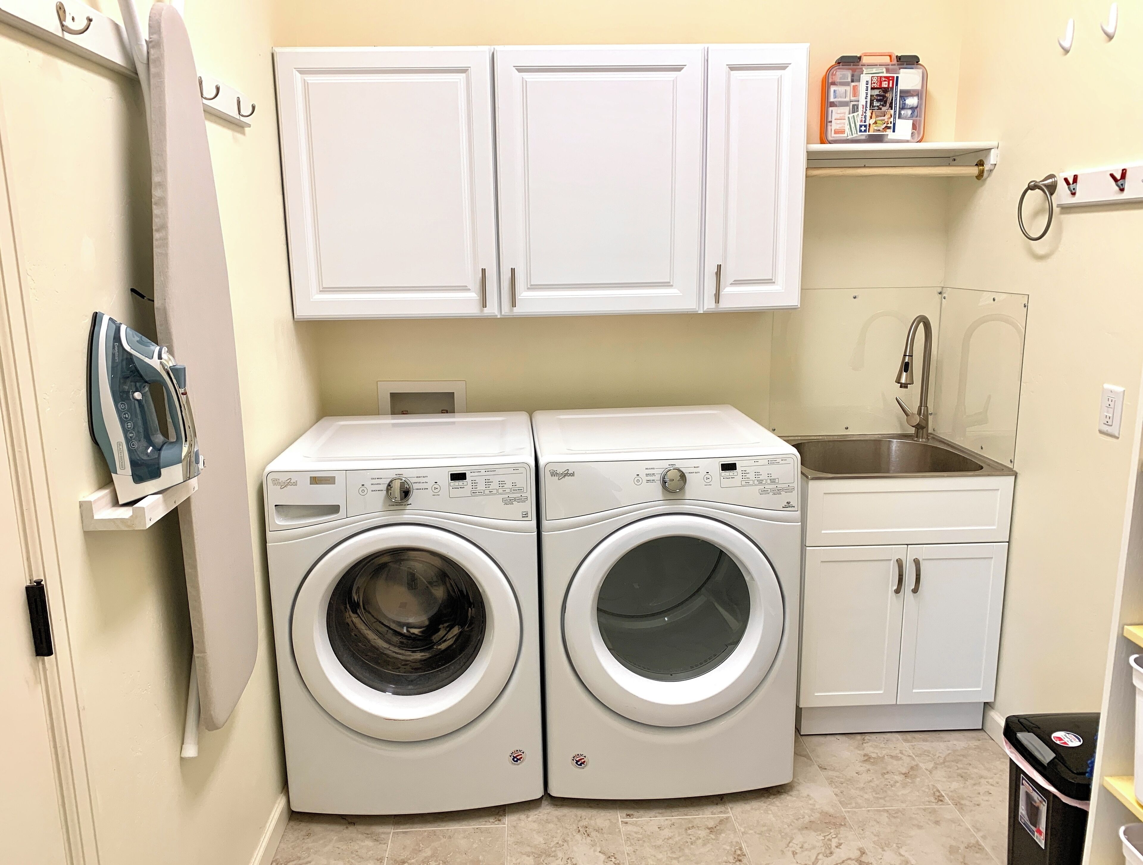 A laundry area with two front-loading washing machines, a drying rack, a small sink, and white cabinets above.