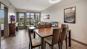 A dining area with a large table and chairs, a TV on a stand, and a view through large windows. - Playa Del Sol Retreat 550 | Pool + Hot Tub | (Kelowna)