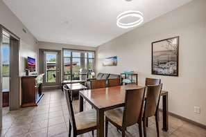 A dining area with a large table and chairs, a TV on a stand, and a view through large windows. - Playa Del Sol Retreat 550 | Pool + Hot Tub | (Kelowna)