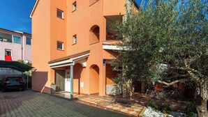 A multi-story building with a checkered pavement in front, a parked car, and a tree with green foliage. - Holiday apartment mit Klimaanlage und Balkon (Krk)