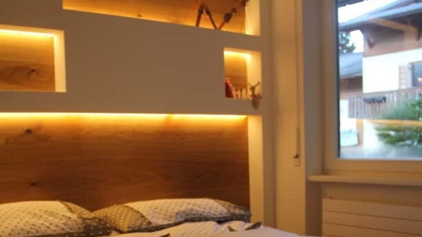 A bedroom with a wooden headboard, a bed with white bedding, and a window with a view of a building. - Fié allo Sciliar (Alpe di Siusi Val Gardena) (Fiè allo Sciliar)