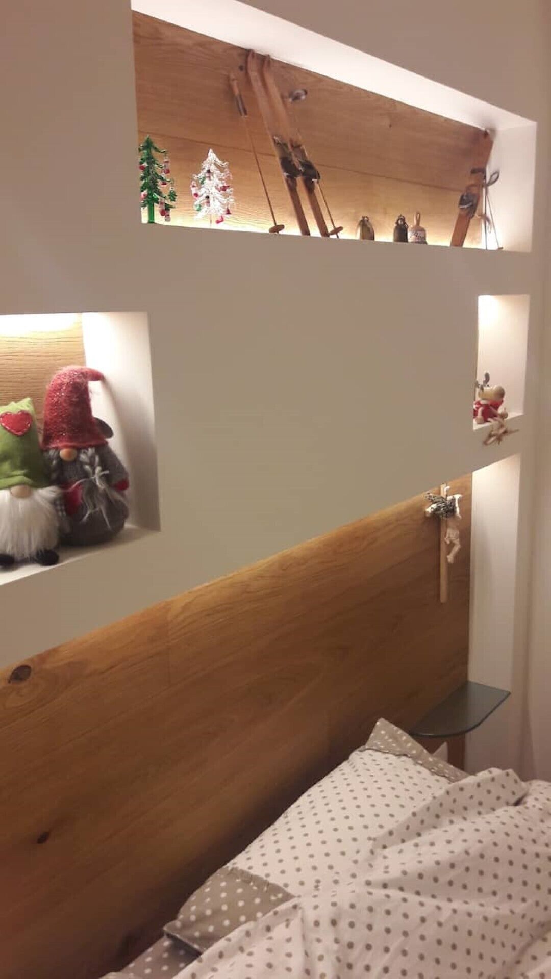 A bedroom with a wooden headboard, a bed with a polka dot bedspread, and shelves above the bed holding various items including a small Christmas tree and figurines.