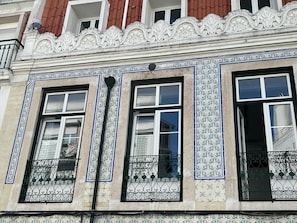 Une façade d’immeuble ornée de carreaux blancs et bleus formant des motifs décoratifs, trois fenêtres à encadrements noirs et des balustrades en métal décoratives.