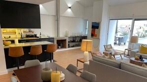 A modern living room with a wooden dining table, grey sofa, and a kitchen area with black cabinets and a built-in TV. - Apart 3Bdr with River view and private Patio - Center of Lisbon (Lisboa)