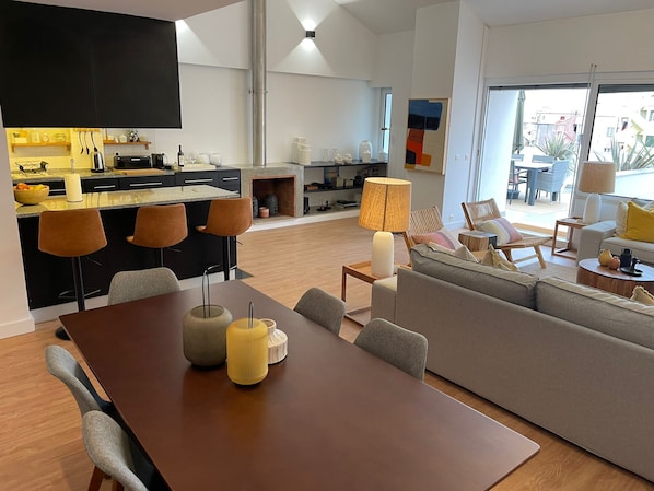 Un salon moderne avec une table à manger en bois, un canapé gris et un coin cuisine doté de meubles noirs et d’un téléviseur encastré.