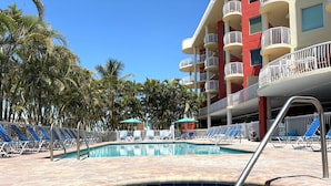 A swimming pool with lounge chairs and umbrellas, surrounded by palm trees and a multi-story building with balconies.