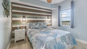 A bedroom with a striped wooden headboard, a bed with a blue and white coral-patterned bedspread, a nightstand with a candle and a small plant, and a window with a view of the outdoors.