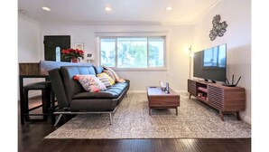 A modern living room with a black leather sofa, a wooden coffee table, a TV stand, and a large window providing natural light.