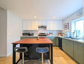 Une cuisine moderne dotée d’un îlot central, de deux tabourets de bar, de meubles blancs et d’une fenêtre laissant entrer la lumière naturelle.