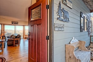 Una puerta de madera con un panel de vidrio, un sofá, una mesa y un banco en una habitación con piso de madera y vistas a montañas a través de ventanas amplias.