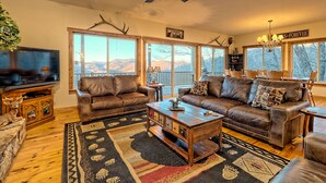 A cozy living room with leather sofas, a wooden coffee table, and a large window offering a mountain view. - Sky's The Limit -Mountain view, golf course, waterfalls, game room. Sleeps 14 (Sky Valley)