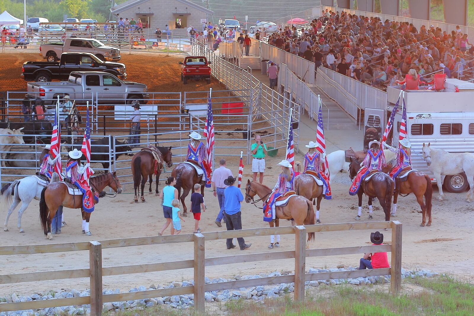 A rodeo event with horses and riders in a fenced arena, spectators in the stands, and a dirt ground.