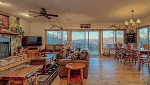 A spacious living room with a fireplace, a large TV, and a dining area with wooden furniture. - Sky's The Limit -Mountain view, golf course, waterfalls, game room. Sleeps 14 (Sky Valley)