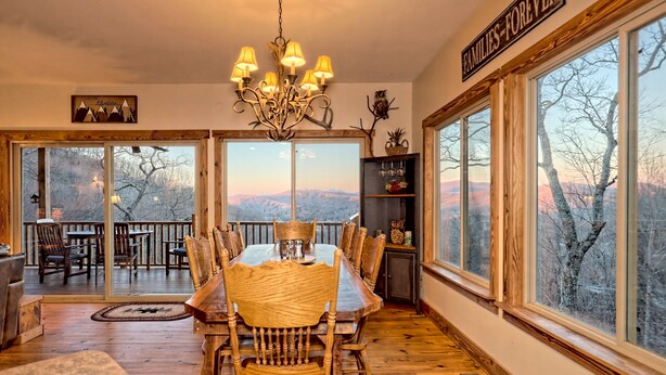 A dining area with a large wooden table, chairs, and a chandelier. There are large windows with a view of mountains. - Sky's The Limit -Mountain view, golf course, waterfalls, game room. Sleeps 14 (Sky Valley)