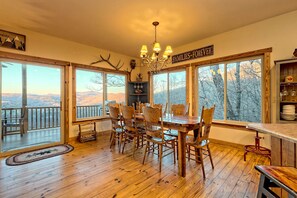 Un comedor con una gran mesa de madera y sillas, una araña y vistas a montañas a través de amplios ventanales.