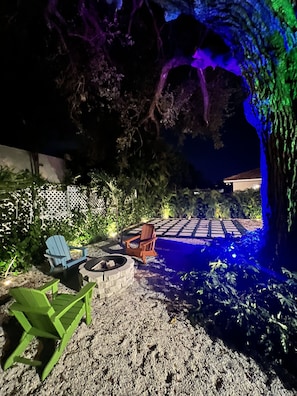 Un patio exterior nocturno con una fogata, dos sillas Adirondack y un pequeño estanque iluminado con luces azules.