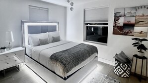 A modern bedroom with a large bed, bedside tables, a nightstand, a window with blinds, a hanging light fixture, and a wall-mounted artwork. - Charming, Cozy in the heart of Miami~Near Airport, Brickell, US1 and the Gables  (Miami)