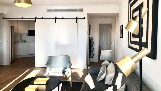 Un salon moderne avec un canapé, une table basse et une lampe suspendue. On aperçoit une cuisine au fond.