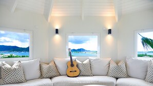 En moderne stue med en hvid sofa, mønstrede puder, en guitar og en bog på et sofabord. Store vinduer giver udsigt over et kystlandskab.