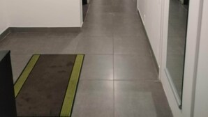 Un couloir étroit doté d’un sol carrelé, d’un luminaire blanc fixé au mur et d’une porte donnant sur une salle de bain.