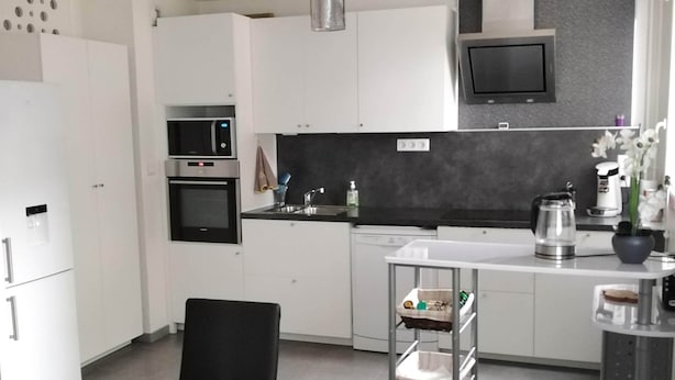 Une cuisine moderne avec des meubles blancs, un plan de travail noir, un four encastré, un four à micro-ondes, une machine à café et une petite table à manger avec des chaises.