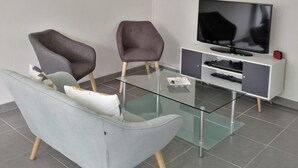 Un salon moderne avec une table basse en verre, deux fauteuils, un téléviseur posé sur un meuble blanc et un petit meuble en dessous.