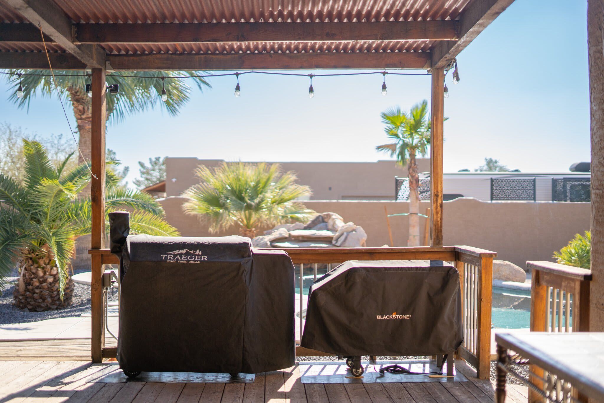 Un espace extérieur abrité, doté de deux housses pour barbecues, l’une portant l’inscription « Traeger » et l’autre « Blackstone », et offrant une vue sur une piscine et des palmiers.