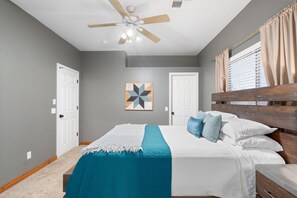 Une chambre à coucher comprenant un lit, un ventilateur de plafond, une fenêtre avec des rideaux et une œuvre d’art accrochée au mur.
