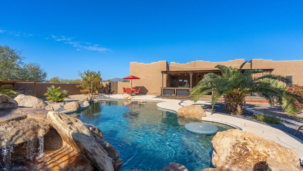A pool area with a clear blue pool, surrounded by rocks and palm trees, and a building with a flat roof in the background. - Western desert escape, pool, backyard oasis (Phoenix)