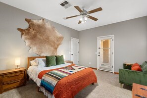 Une chambre avec un grand lit, un ventilateur de plafond, une table de chevet, un fauteuil vert et une porte avec des stores.