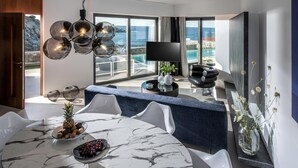 A modern dining area with a marble table, white chairs, and a blue cushioned bench. A large TV is mounted on the wall, and the room has large windows with a view of the sea. - Made By The Sea Villas 8 Bdrm (Malevizi)