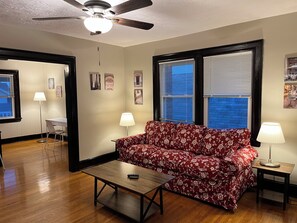 Uma sala de estar com sofá, mesa de centro, ventilador de teto e janela com persianas.