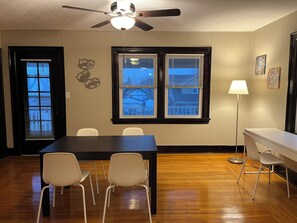 Quarto com piso de madeira, mesa de jantar escura com quatro cadeiras, ventilador de teto e duas janelas com persianas.