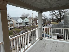 A covered porch with a white railing, overlooking a residential neighborhood with houses and parked cars.