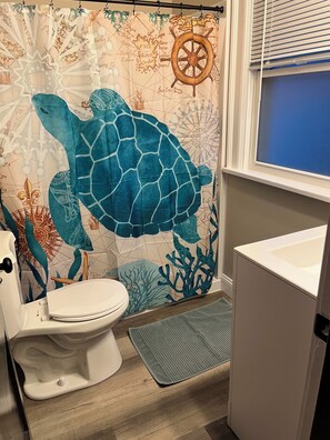 Um banheiro com cortina de chuveiro com tema de tartaruga marinha, vaso sanitário branco e janela com persianas.